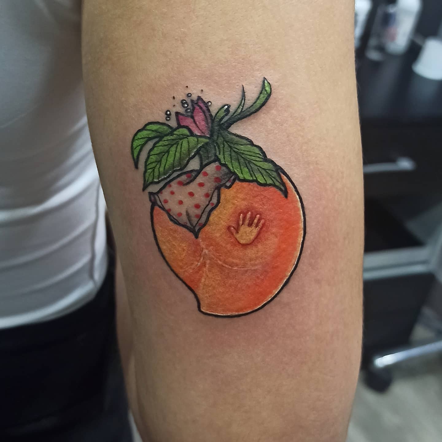 A tattoo of a peach with a mini hand and a strawberry wearing a flower crown on an arm